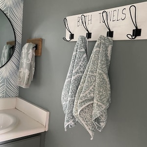 May include: White wooden bathroom towel rack with black metal hooks. The rack has the words "BATH TOWELS" written on it. Two blue and white patterned towels are hanging on the hooks.