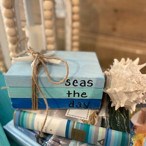 May include: A stack of three wooden blocks painted in shades of blue, tied with twine. The top block has the words "Seas the day" painted in black. The blocks are sitting on top of a stack of books with blue and white striped covers.