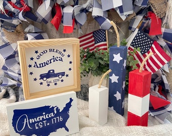 May include: Three wooden signs with patriotic themes. The first sign is a light brown wooden frame with a white background and the text "God Bless America Est. 1776" in blue. The second sign is a white wooden sign with a blue outline of the United States and the text "America EST 1776" in white. The third sign is a blue wooden sign with white stars and the text "God Bless America" in white. The signs are surrounded by red, white, and blue fabric.