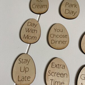Op de afbeelding: Houten, eivormige tokens met verschillende activiteiten erop geschreven, waaronder "Ijs", "Dagje uit in het park", "Dag met mama", "Jij kiest het diner", "Laat opblijven", "Extra schermtijd" en "Spelletjesavond".