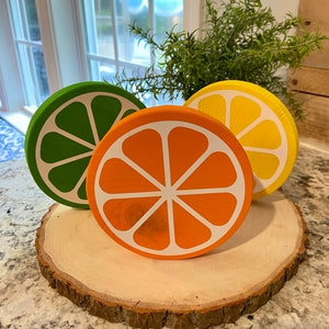 Puede incluir: Tres rodajas de fruta c&iacute;trica de madera sobre una rodaja de madera. Las rodajas est&aacute;n pintadas en tonos de verde, naranja y amarillo. Las rodajas de fruta est&aacute;n dispuestas en c&iacute;rculo sobre la rodaja de madera.
