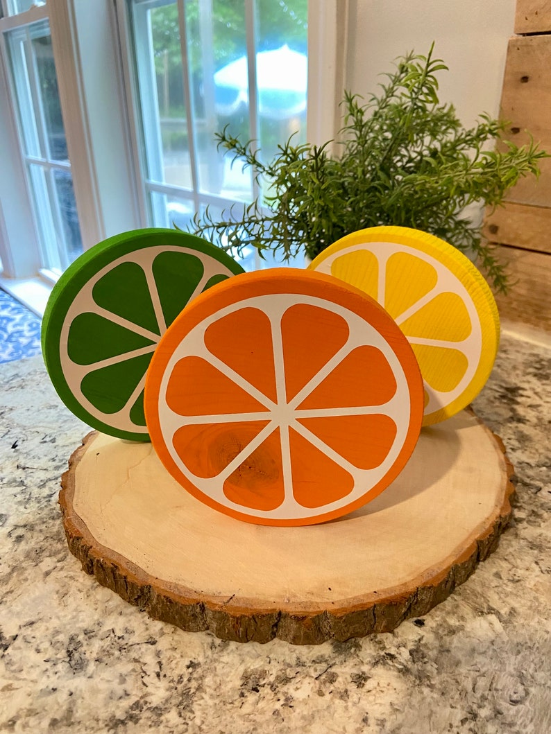 Puede incluir: Tres rodajas de fruta c&iacute;trica de madera sobre una rodaja de madera. Las rodajas est&aacute;n pintadas en tonos de verde, naranja y amarillo. Las rodajas de fruta est&aacute;n dispuestas en c&iacute;rculo sobre la rodaja de madera.