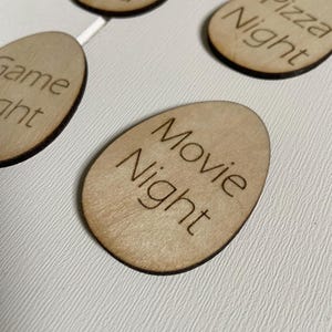 Op de afbeelding: Houten eivormige ornamenten met verschillende activiteiten erop geschreven, waaronder "Filmavond", "Pizza-avond" en "Dag met papa".