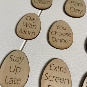 Op de afbeelding: Houten, eivormige tokens met verschillende activiteiten erop geschreven, waaronder "IJs", "Dagje uit in het park", "Dag met mama", "Jij kiest het diner", "Laat opblijven", "Extra schermtijd".