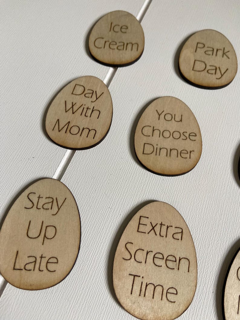 Op de afbeelding: Houten, eivormige tokens met verschillende activiteiten erop geschreven, waaronder "IJs", "Dagje uit in het park", "Dag met mama", "Jij kiest het diner", "Laat opblijven", "Extra schermtijd".
