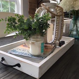 May include: A white wooden tray with black handles holds a decorative arrangement. It features a white lantern with wooden beads, a candle, greenery, and magazines. A glass jar with a candle and a bouquet of flowers are also present.