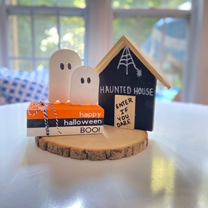 May include: A wooden haunted house with a black sign that reads "Haunted House Enter If You Dare" and a spider web on the roof. Two white ghosts are in front of the house. A small orange and white box with the text "Happy Halloween Boo!" is in front of the house.