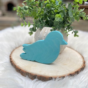 May include: A light blue wooden bird figurine on a wooden slice. The bird is sitting on a white fuzzy surface.