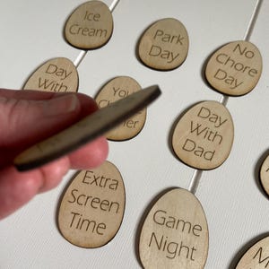 Op de afbeelding: Houten, eivormige tokens met verschillende activiteiten erop geschreven, zoals "IJs", "Dagje uit", "Geen klusjesdag", "Dag met papa", "Extra schermtijd", "Spelletjesavond".