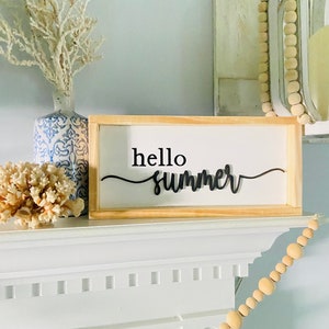 Op de afbeelding: Een houten bord met de tekst "hello summer" in zwarte letters op een witte achtergrond. Het bord staat op een witte schouw met een witte vaas en een koraaltak links en een houten kralensnoer rechts.