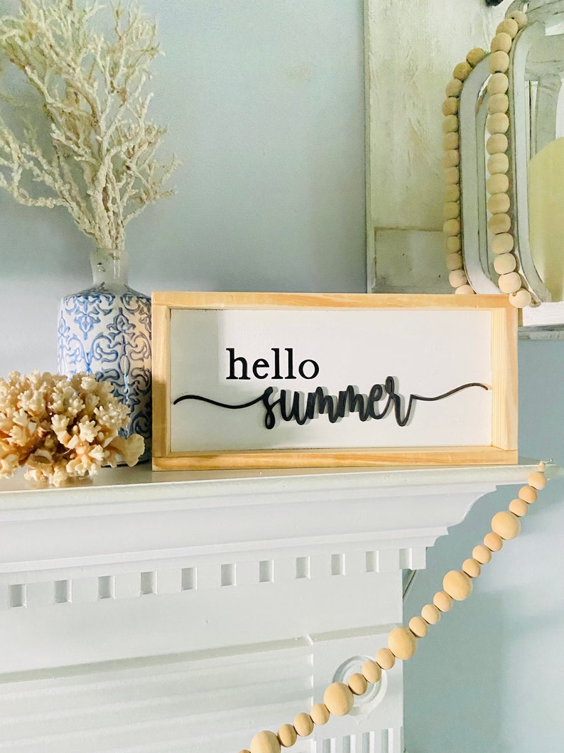 Op de afbeelding: Een houten bord met de tekst "hello summer" in zwarte letters op een witte achtergrond. Het bord staat op een witte schouw met een witte vaas en een koraaltak links en een houten kralensnoer rechts.