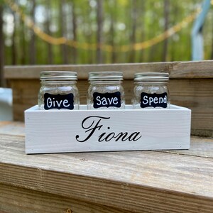May include: White wooden box with three glass jars labeled "Give", "Save", and "Spend". The box is labeled "Fiona".