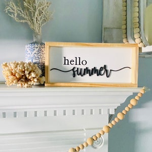 Op de afbeelding: Een houten bord met de tekst "hello summer" in zwarte cursieve letters. Het bord staat op een witte schoorsteenmantel met een witte koraal en een blauw-witte vaas met een witte tak.