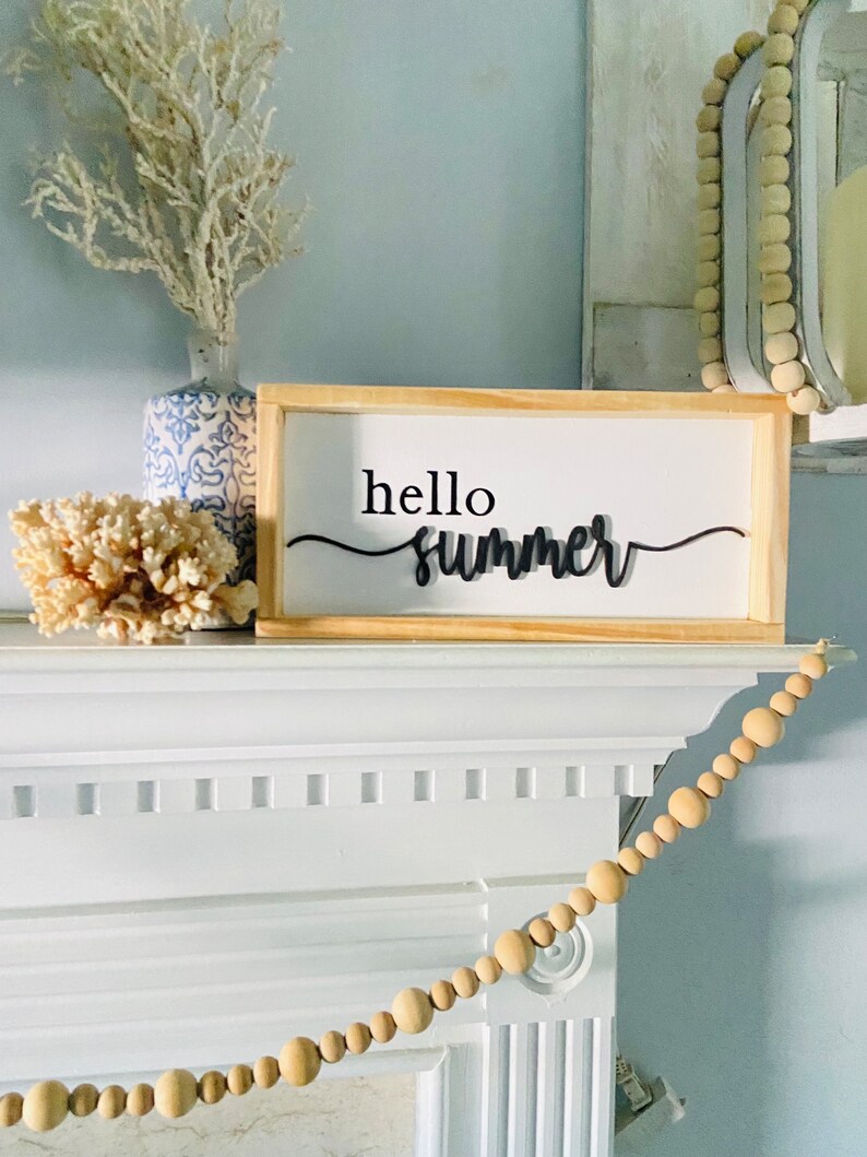 Op de afbeelding: Een houten bord met de tekst "hello summer" in zwarte cursieve letters. Het bord staat op een witte schoorsteenmantel met een witte koraal en een blauw-witte vaas met een witte tak.