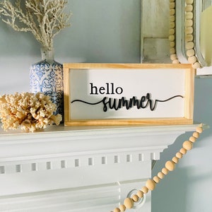Op de afbeelding: Een houten bord met de tekst "hello summer" in zwarte cursieve letters. Het bord staat op een witte schouw met een houten kralenslinger eroverheen.