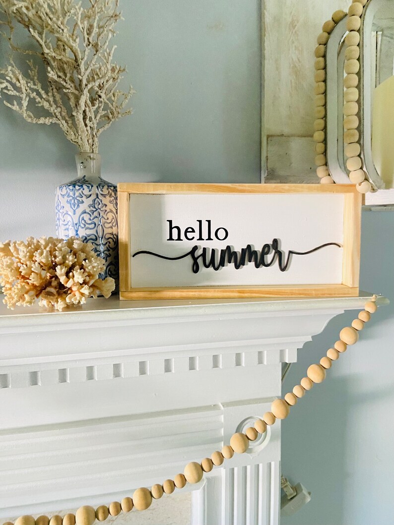 Op de afbeelding: Een houten bord met de tekst "hello summer" in zwarte cursieve letters. Het bord staat op een witte schouw met een houten kralenslinger eroverheen.