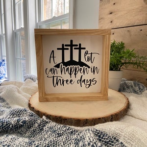 Puede incluir: Un letrero de madera con fondo blanco y letras negras. El letrero dice "A lot can happen in three days". El letrero est&aacute; decorado con tres cruces negras.