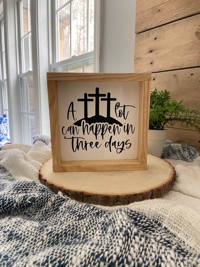 Puede incluir: Un letrero de madera con fondo blanco y letras negras. El letrero dice "A lot can happen in three days". El letrero est&aacute; decorado con tres cruces negras.