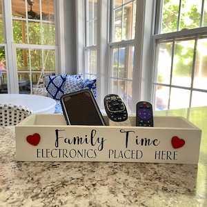 May include: White wooden box with the text "Family Time Electronics Placed Here" and two red hearts. The box contains a cell phone, a remote control, and a second remote control.