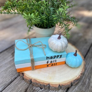 Puede incluir: Una pila de tres libros de madera pintados con las palabras "happy fall y'all" en la parte delantera. Los libros est&aacute;n sobre una rodaja de madera con una calabaza blanca y una azul a su lado.