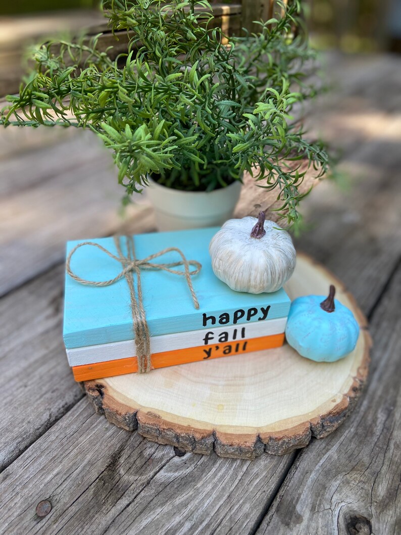 Puede incluir: Una pila de tres libros de madera pintados con las palabras "happy fall y'all" en la parte delantera. Los libros est&aacute;n sobre una rodaja de madera con una calabaza blanca y una azul a su lado.