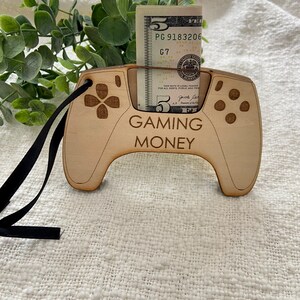 May include: A wooden cutout of a video game controller with a slot for a five dollar bill. The controller is painted brown and has the words "GAMING MONEY" printed on it.