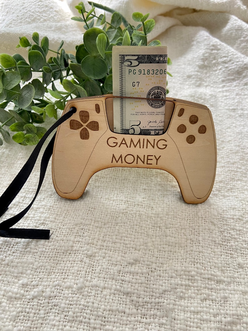 May include: A wooden cutout of a video game controller with a slot for a five dollar bill. The controller is painted brown and has the words "GAMING MONEY" printed on it.