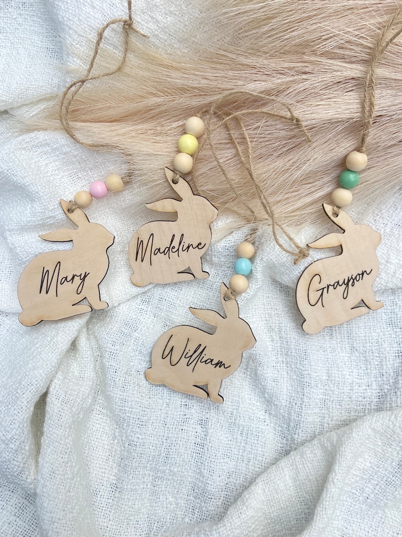 K&ouml;nnte beinhalten: Vier h&ouml;lzerne Hasen-Anh&auml;nger mit den Namen Mary, Madeline, William und Grayson darauf geschrieben. Jeder Hase ist an einer Schnur mit Holzperlen in verschiedenen Farben befestigt.