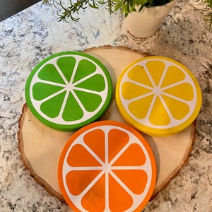 Puede incluir: Tres posavasos de madera con forma de rodajas de c&iacute;tricos. Los posavasos est&aacute;n pintados de verde, amarillo y naranja, con contornos blancos. Los posavasos est&aacute;n dispuestos sobre una rodaja de madera.