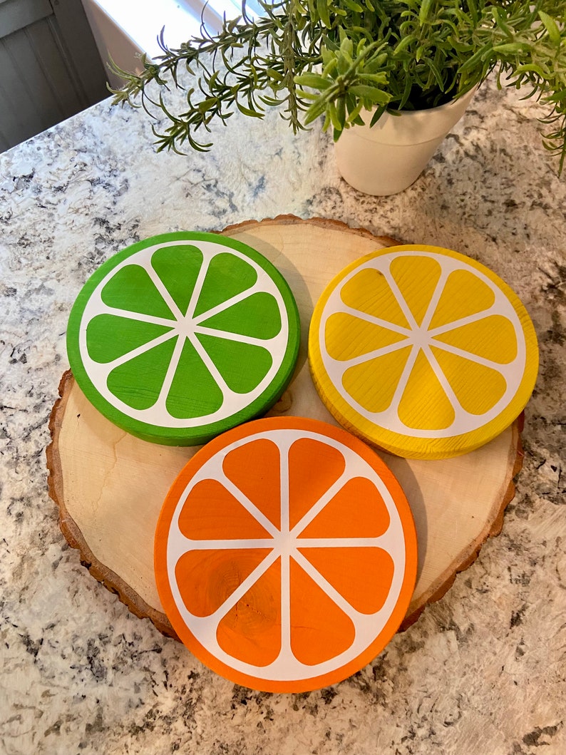 Puede incluir: Tres posavasos de madera con forma de rodajas de c&iacute;tricos. Los posavasos est&aacute;n pintados de verde, amarillo y naranja, con contornos blancos. Los posavasos est&aacute;n dispuestos sobre una rodaja de madera.