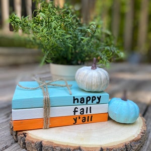 Puede incluir: Tres bloques de madera apilados sobre una rodaja de madera. Los bloques est&aacute;n pintados de azul, blanco y naranja. El bloque superior tiene las palabras "happy fall y'all" escritas en negro. Dos peque&ntilde;as calabazas blancas y azules est&aacute;n a un lado.
