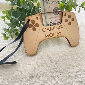 May include: A wooden cutout of a video game controller with a slot for a dollar bill. The controller is painted brown and has the words "GAMING MONEY" printed on it. A five dollar bill is inserted into the slot.