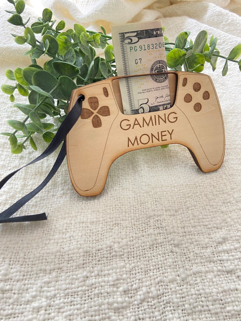 May include: A wooden cutout of a video game controller with a slot for a dollar bill. The controller is painted brown and has the words "GAMING MONEY" printed on it. A five dollar bill is inserted into the slot.