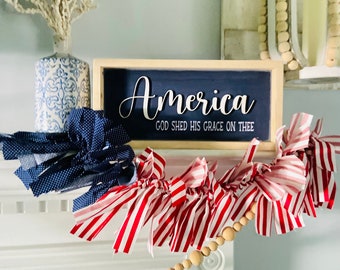 May include: A wooden sign with the word "America" in white cursive lettering and the phrase "God Shed His Grace On Thee" in smaller white lettering. The sign is on a white mantel with a red and white striped fabric garland and a wooden bead garland.