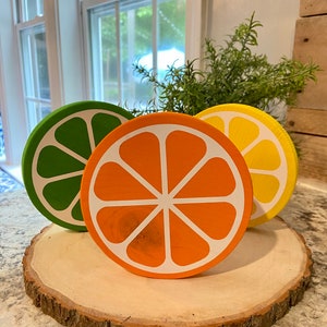 Puede incluir: Tres rodajas de fruta de madera sobre una rodaja de madera. Las rodajas est&aacute;n pintadas para parecer un lim&oacute;n verde, una naranja y un lim&oacute;n. El lim&oacute;n verde es verde, la naranja es naranja y el lim&oacute;n es amarillo.