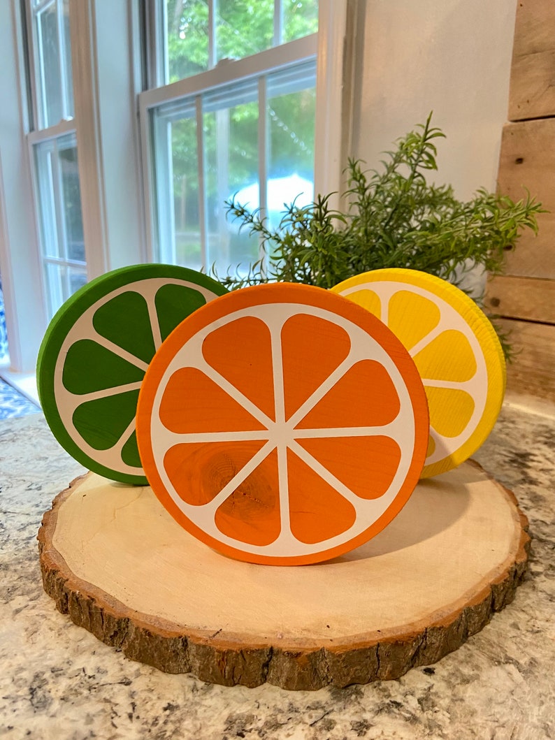 Puede incluir: Tres rodajas de fruta de madera sobre una rodaja de madera. Las rodajas est&aacute;n pintadas para parecer un lim&oacute;n verde, una naranja y un lim&oacute;n. El lim&oacute;n verde es verde, la naranja es naranja y el lim&oacute;n es amarillo.