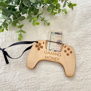 May include: A wooden video game controller ornament with a slot for cash. The ornament is brown and has the text "Gaming Money" on it. A five dollar bill is inserted into the slot.