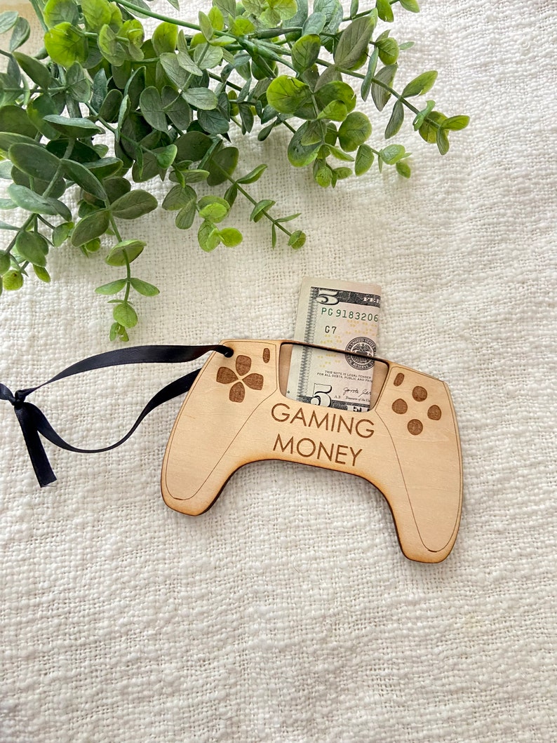 May include: A wooden video game controller ornament with a slot for cash. The ornament is brown and has the text "Gaming Money" on it. A five dollar bill is inserted into the slot.