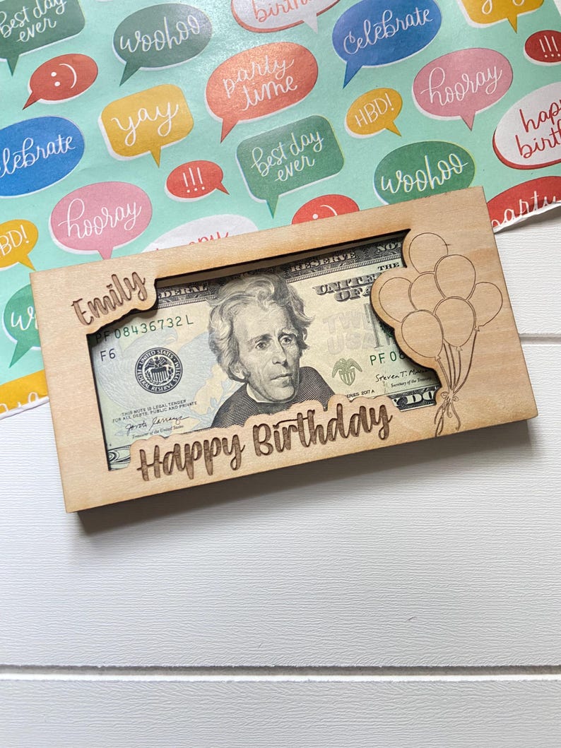 May include: A wooden money holder engraved with "Happy Birthday" and "Emily". It displays a twenty-dollar bill through a cutout. A laser-cut balloon design is on the right. The background is patterned with colourful speech bubbles, ideal for a birthday gift.