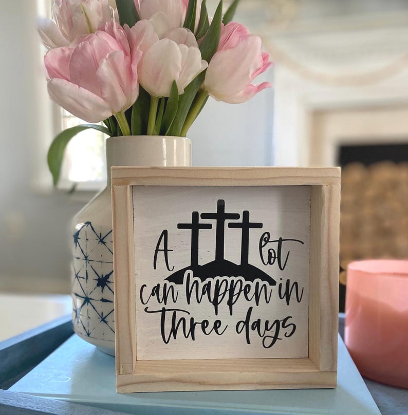 Puede incluir: Un letrero de madera con fondo blanco y letras negras. El letrero dice "A lot can happen in three days". Hay tres cruces negras en el letrero, que representan la crucifixi&oacute;n de Jesucristo. El letrero est&aacute; sobre una superficie azul con flores rosas en un jarr&oacute;n detr&aacute;s.