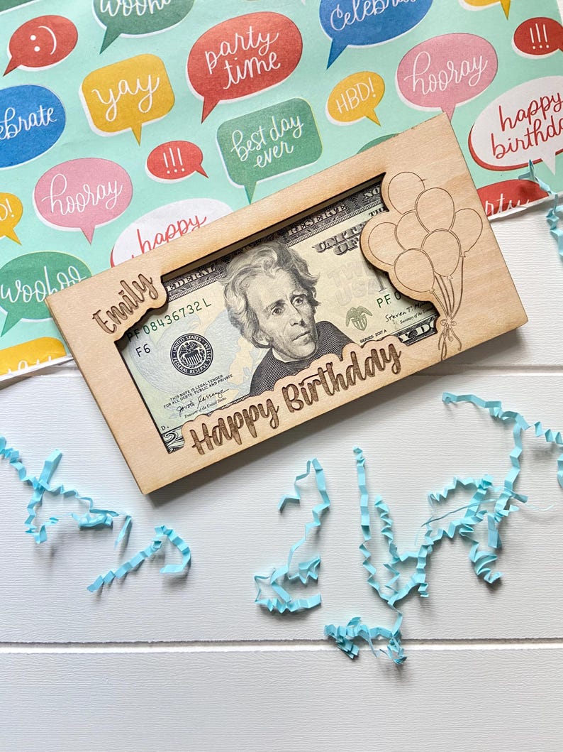 May include: A wooden money holder engraved with "Emily" and "Happy Birthday". It frames a twenty-dollar bill, with a balloon design. The background features colourful speech bubbles and blue paper streamers.
