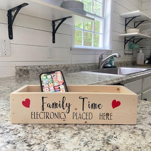 May include: A rectangular light-colored wooden storage box featuring the black text "Family Time" and "ELECTRONICS PLACED HERE" with a small red heart on each side. A smartphone is resting inside the box, which is positioned on a countertop.