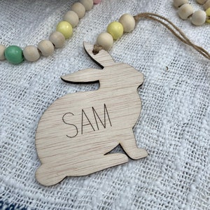 K&ouml;nnte beinhalten: Ein ausgeschnittener Holzhase mit dem eingravierten Namen "SAM". Der Hase h&auml;ngt an einer Schnur mit pastellfarbenen Holzperlen.