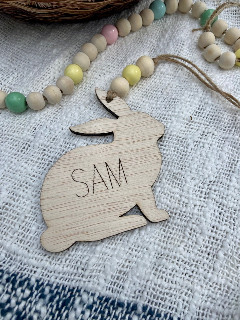 K&ouml;nnte beinhalten: Ein ausgeschnittener Holzhase mit dem eingravierten Namen "SAM". Der Hase h&auml;ngt an einer Schnur mit pastellfarbenen Holzperlen.