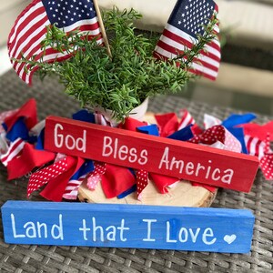 May include: Two wooden signs with patriotic messages. The top sign is red and says "God Bless America". The bottom sign is blue and says "Land that I Love". The signs are surrounded by red, white, and blue fabric scraps and two small American flags.