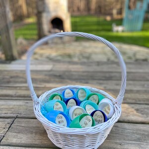 Op de afbeelding: Een witte rieten mand gevuld met blauwe en groene plastic flesdoppen. Elke dop heeft een ander woord erop geschreven, zoals "Dag", "Geen klusje", "Koekjes bakken", "IJs", "Extra portie", "Film", "Spel", "Blijven", "Park", "Pizza avond", "Jij kiest".