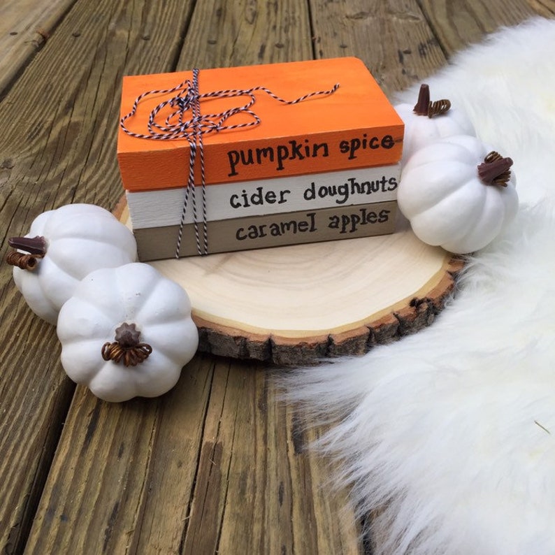 K&ouml;nnte beinhalten: Drei gestapelte Holzb&uuml;cher mit Herbstmotiven: "pumpkin spice", "cider doughnuts" und "caramel apples". Die B&uuml;cher sind mit orangefarbenem und wei&szlig;em Garn zusammengebunden und liegen auf einer Holzscheibe mit wei&szlig;em Kunstfell.