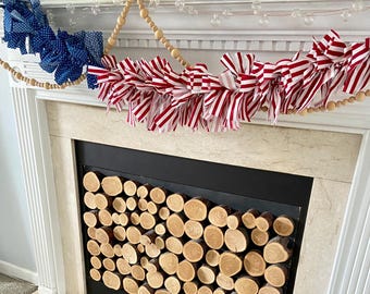 May include: A red, white, and blue fabric garland with a wooden bead garland draped over it, hanging above a fireplace with a stacked log fire screen.