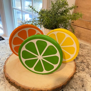 Puede incluir: Tres rodajas de fruta de madera, pintadas para parecer una naranja, una lima y un lim&oacute;n, est&aacute;n dispuestas sobre una rodaja de madera. Las rodajas de fruta est&aacute;n pintadas en colores brillantes y realistas.
