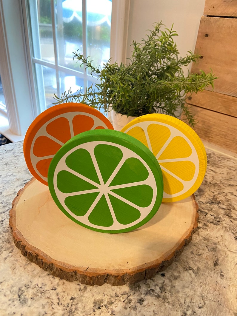 Puede incluir: Tres rodajas de fruta de madera, pintadas para parecer una naranja, una lima y un lim&oacute;n, est&aacute;n dispuestas sobre una rodaja de madera. Las rodajas de fruta est&aacute;n pintadas en colores brillantes y realistas.